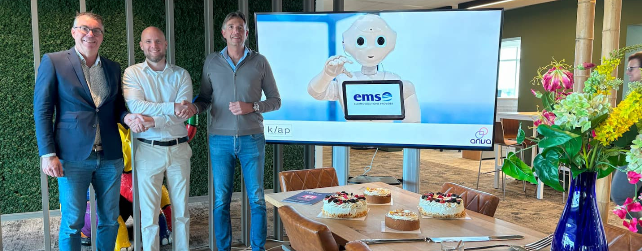 Drie vertegenwoordigers van Klap, EMS en ANVA schudden elkaar de hand voor een scherm met de EMS-robot en logo’s; op tafel staan taarten in een moderne kantoorruimte.