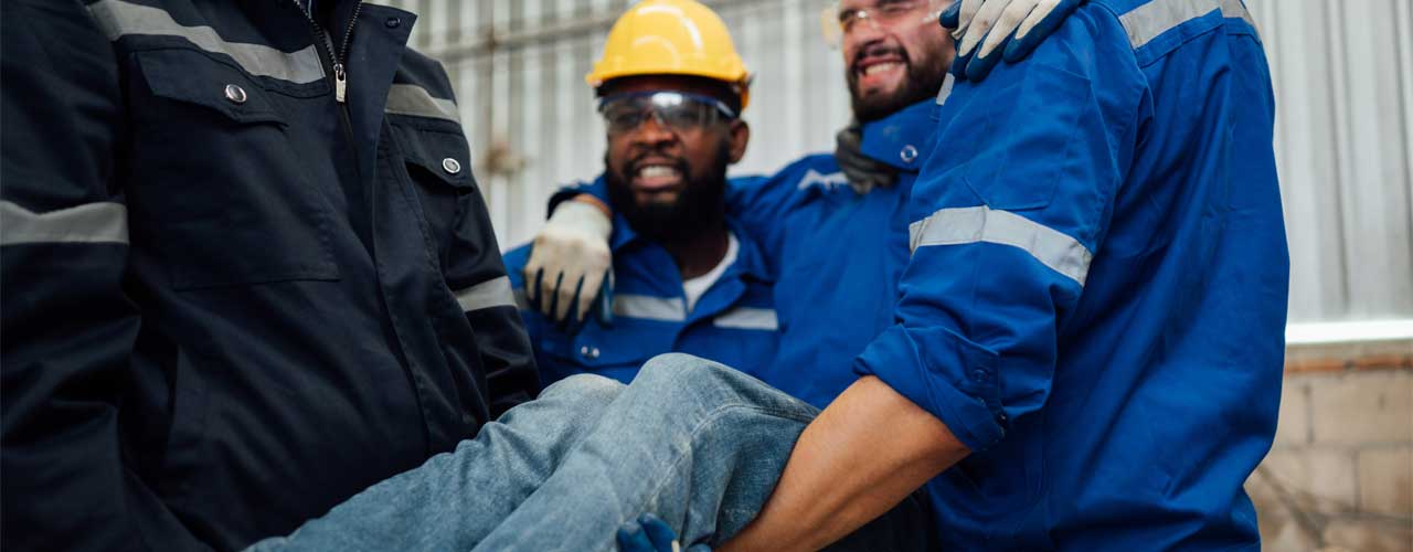 Twee werknemers in werkoveralls dragen een geblesseerde collega tijdens een bedrijfsongeval; iedereen draagt persoonlijke beschermingsmiddelen.